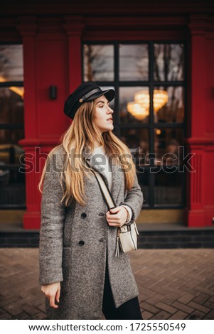 a stylish girl on a background a beautiful red facade is dressed in a grey overcoat jeans leather shoe on a heel and with a head-dress by a hat by a cap and white bag, street style fashion