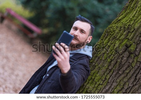 A handsome young hipster man leaning against a tree and taking a selfie in the park.