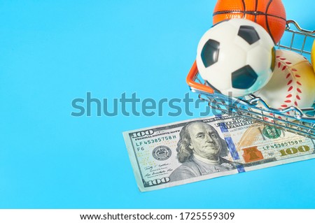 Different balls and banknote of 100 dollars in metal market basket on blue background. Purchasing sport accessories. Concept of corruption in sport