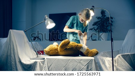 Cute little girl wearing doctor suit playing at home, pretending to be a surgeon, bandaging her teddy bear Royalty-Free Stock Photo #1725563971