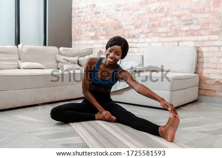 African american sportswoman smiling, looking at camera and stretching on yoga mat in living room