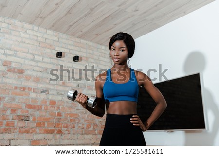 African american sportswoman with hand on hip training with dumbbell and looking at camera in living room