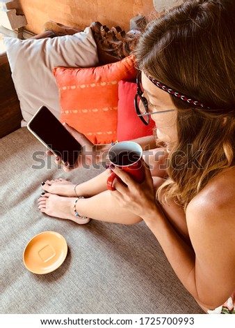 Modern young woman using cellphone and drinking coffee on a terrace sofa.