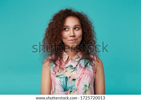 Portrait of charming young curly brunette woman with natural makeup biting her lips while looking positively aside, isolated over blue background with hands down