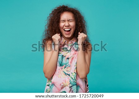 Overjoyed young attractive curly brunette woman raising emotionally her hands while rejoicing about something with closed eyes, isolated over blue background