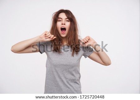 Drowsy young lovely brunette woman with casual hairstyle raising her hands while stretching herself and keeping mouth opened while yawning, isolated over white background