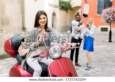 Pretty cheerful brunette Caucasian girl sitting on the red scooter in old city street, while her multiracial friends, African guy and Caucasian girl having fun and making selfie photo