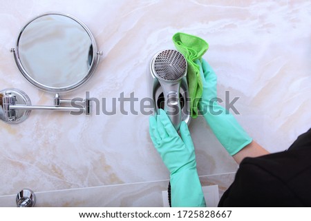A woman in a cleaning service uniform wipes a hair dryer in the bathroom. Unrecognizable photo. The concept of cleanliness and hygiene.