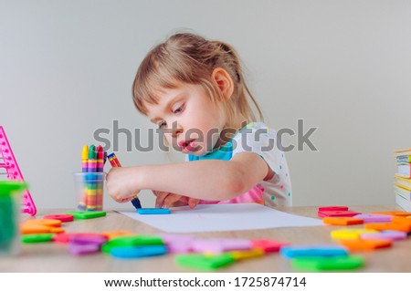 Beautiful little girl drawing with wax crayon and blue square shape sitting at the table. Grey wall as a bckground with blank space for text.

