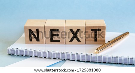 Next Word Written In Wooden Cubes on white notebook and blue table