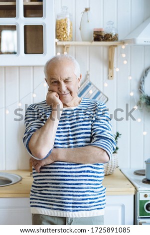 Man aged, grandfather in cheerful mood winks while looking at camera on background of kitchen. Retirement age, old people and age category of people.