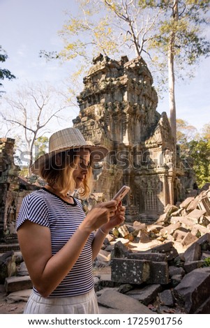 A young woman wearing a hat is checking her phone in Angkor Temple ruins.