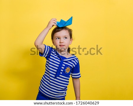 A cute little girl in a sailor suit holds a paper boat on her head and looks up at the yellow background. The day of the Navy.