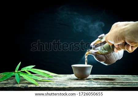 pouring hot tea from a teapot into cup steam is over the cupping, bamboo leaves,  on the old wood table a black background Left  side copy space