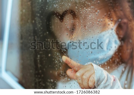 Little girl in face mask drawing heart symbol on the window glass with rain drops. Selective focus on the heart. Social isolation stay at home during Pandemic COVID-19 concept.