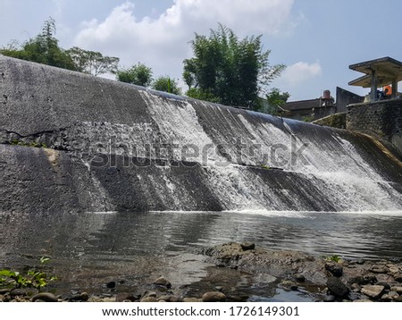Photography of waterfall from the top of the Yellow River Dam in Sleman, Indonesia. The water runs through fish ponds and surrounding rice fields