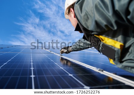 Electrician in safety helmet, uniform and working gloves, setting a shiny new solar battery with help of hex key, blue sky on background. Concept of alternative green energy sources and innovations. Royalty-Free Stock Photo #1726252276