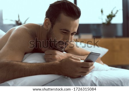 Smiling handsome man is lying on pillow and messaging on smartphone soon after waking up stock photo