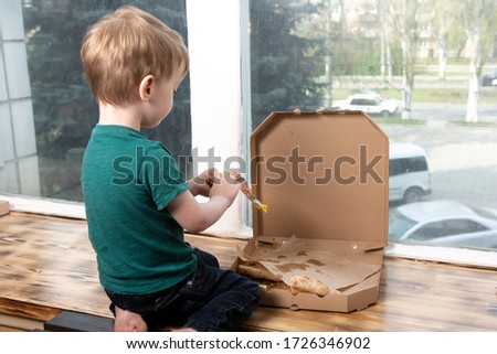 a three year old child eats a slice of pizza from a box. the boy is turned his back and looks out the window into the street. quarantined leisure time. delivery of food from a pizzeria.