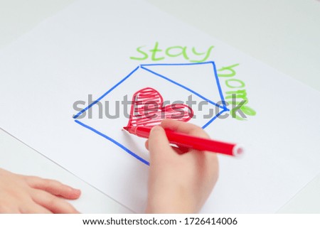 Close up of hand of child drawing house with red heart on the sheet of paper with a painted words Stay Home over the house at home. Stay Home concept. Selective focus.