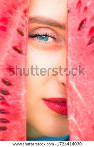 Eye of young woman peeks between two slices of watermelon.