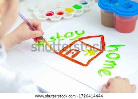 Child is drawing house and Stay Home words by brush with watercolors on white paper at home.