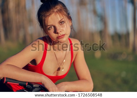 Cute girl with pendant on a chain in a red top with her hair tied in a bun, leaned on the handlebars of a bicycle. sunset on nature against the background of the forest and trees. Close-up.