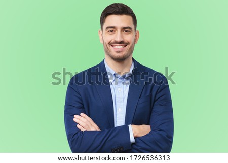 Handsome young man in formal blazer, standing in front of camera with positive confident smile, holding arms crossed, feeling relaxed, isolated on green background