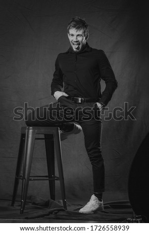 Young bearded man grimacing in studio wearing black shirt and trousers and white gumshoes. Black and white photo.