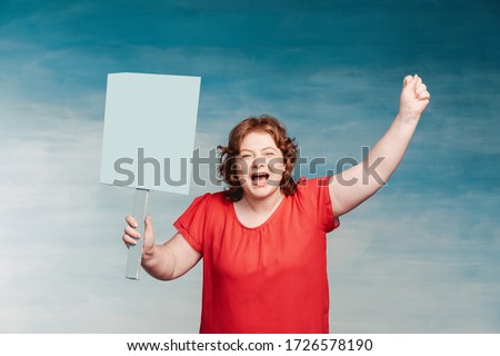Cute smiling chubby red-haired woman screaming fervently, waving his hand in the second holds a protest poster on a blue background.