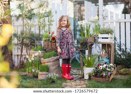 Cute little curly blonde toddler girl in red rubber boots posing in the garden among flowers