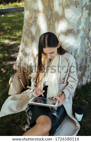 under a huge plane tree in the park, a girl sits with a tablet pc in her hands and writes something with her finger on the screen