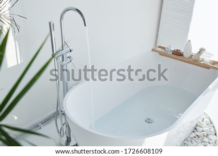 White bathroom modern interior. Luxurious decor with plants, window, spa at home. Bathtub is filled with tap water, nobody Royalty-Free Stock Photo #1726608109