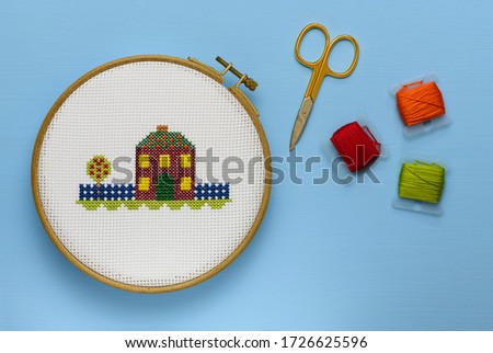 The cross-stitch fall house in the wooden hoop, one metal scissors and three plastic bobbin with the colored threads are on the blue background, the view from above.
