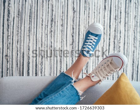 Relaxing Teenager's feet in different colors beige and blue casual new sneakers on cozy comfortable living room sofa over striped design mat. Comfortable clothing and footwear concept top view image.