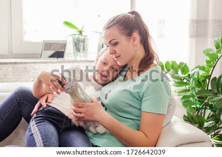 Mother with her adult daughter sitting together and having good time in the living room in front of window. Daughter shows mom a doll