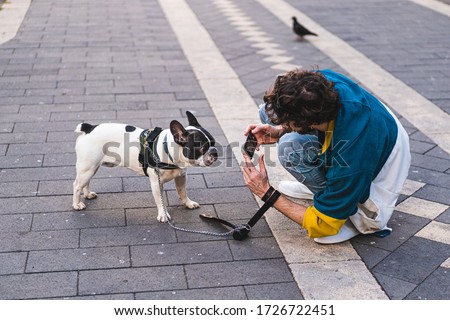 Young man taking picture of his puppy dog outdoor with the smartphone. French bulldog posing for pic shot for his owner.