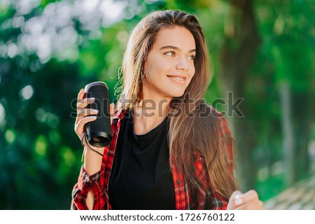 Modern trendy girl listening to music by wireless portable speaker.Young beautiful american woman enjoying,dancing in park.