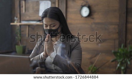 Asian Businesswoman Prays In Office Putting Her Palms Together With Closed Eyes While Sitting At Table, Pandemic Concept.