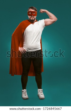 Super Hero Man Shows His Muscles And Ready To Save World. Serious Man In Red Super Hero Mask And Coat Showing His Muscles Looking At Camera. Cut Out On Biscay Green Background.