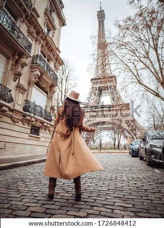 A stylish girl in a beige coat, hat and boots stands on a small Parisian street with a view of the Eiffel Tower. Paris style. Royalty-Free Stock Photo #1726848412