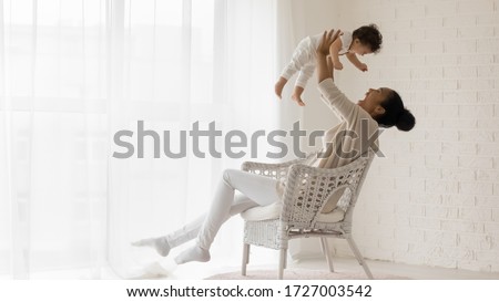 Happy loving african American young mother sit in chair at home relax play with funny little newborn baby girl, smiling biracial mom have fun lift in air enjoy family weekend with cute infant child Royalty-Free Stock Photo #1727003542