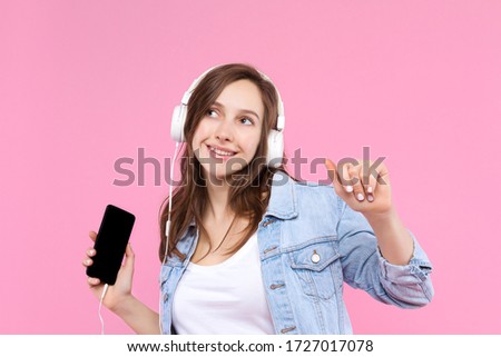 Pretty, smiling girl is holding in hand smartphone and listening music. Young woman dressed in denim jacket, white t-shirt, headphones on pink background. Modern technology and devices concept.