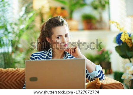 pensive trendy middle age housewife in blue blouse and striped jacket sitting on divan with laptop in the modern house in sunny day.