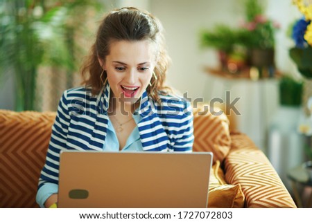 surprised modern woman in blue blouse and striped jacket sitting on couch with laptop in the modern house in sunny day.