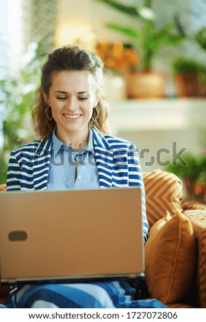 happy trendy woman in blue blouse and striped jacket sitting on couch with laptop in the modern house in sunny day.