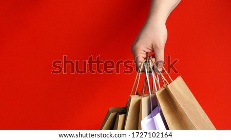 Hand with white paper bags and craft on bright red background. Season of sales or purchases as gift for holidays. Christmas shopping. Courier delivery of products in eco-friendly packaging Royalty-Free Stock Photo #1727102164