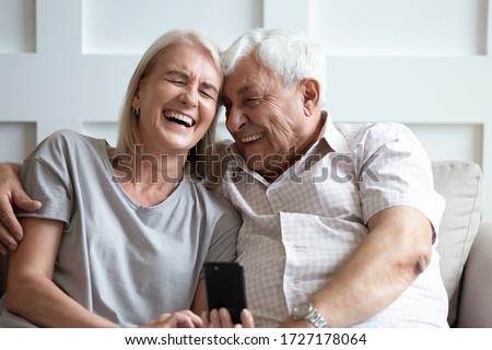 Overjoyed mature couple relax on couch have fun watching funny video on cellphone together, happy senior husband and wife sit rest on sofa at home laugh smile using modern smartphone