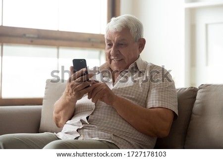 Smiling mature 80s man sit on sofa in living room texting messaging on modern smartphone gadget, happy senior 70s grandfather rest on couch at home browse internet on cellphone, technology concept Royalty-Free Stock Photo #1727178103