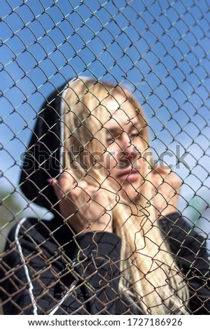 A girl with white hair in a hood stands behind a metal chain-link fence on the Park's Playground. Blurry photo